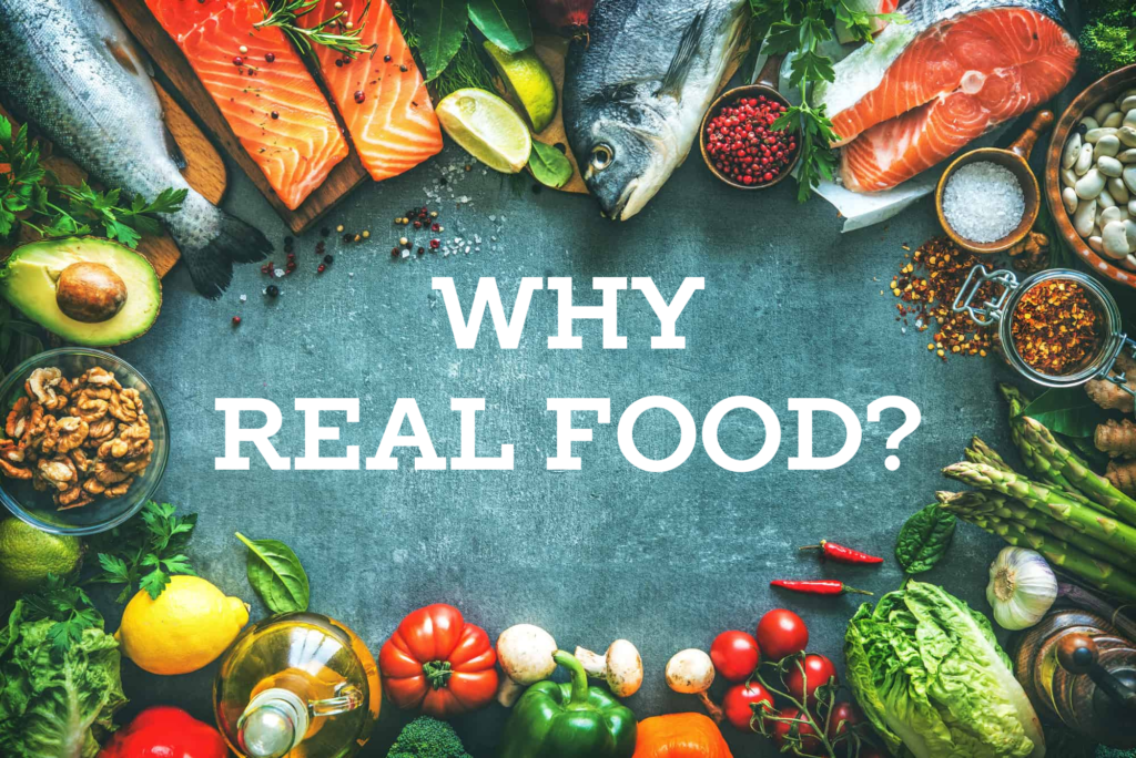 A variety of fresh and healthy foods arranged in a border around a dark surface, including whole fish, salmon fillets, avocado, walnuts, peppers, tomatoes, asparagus, herbs, spices, and other colorful vegetables. The text "Why Real Food?" is prominently displayed in the center.