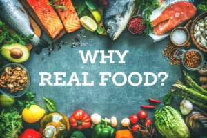 A variety of fresh and healthy foods arranged in a border around a dark surface, including whole fish, salmon fillets, avocado, walnuts, peppers, tomatoes, asparagus, herbs, spices, and other colorful vegetables. The text "WHY REAL FOOD?" is prominently displayed in the center.